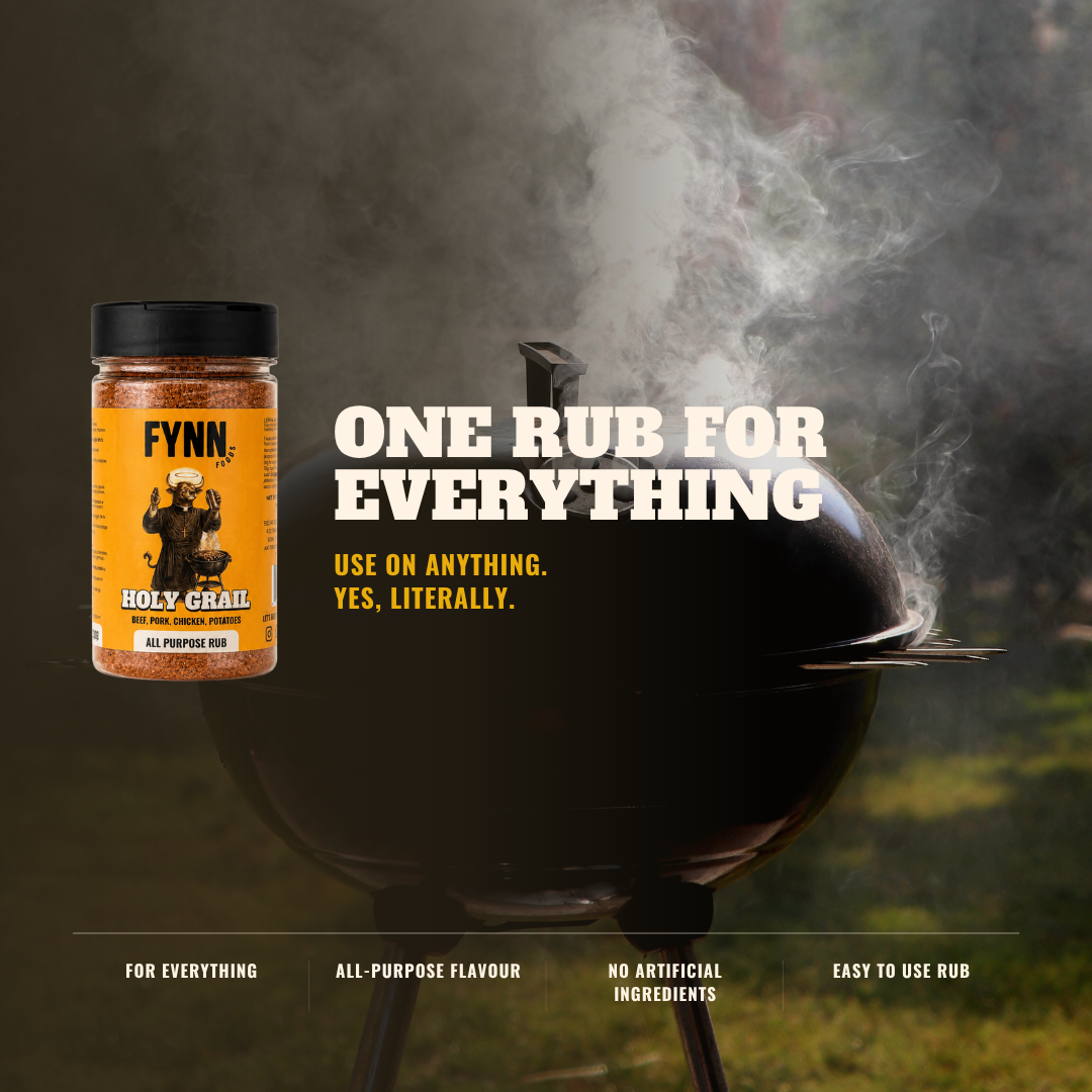 A jar of Fynn Holy Grail All Purpose Rub shown next to a black kettle grill with smoke rising in an outdoor setting. The product label is yellow and black, and the image features marketing text that reads 'ONE RUB FOR EVERYTHING' and 'USE ON ANYTHING.